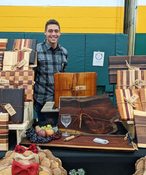 A man in a flannel shirt smiles as he shows off his woodworking items at the Franklin D. Roosevelt High School Holiday Craft Fair in Staatsburg